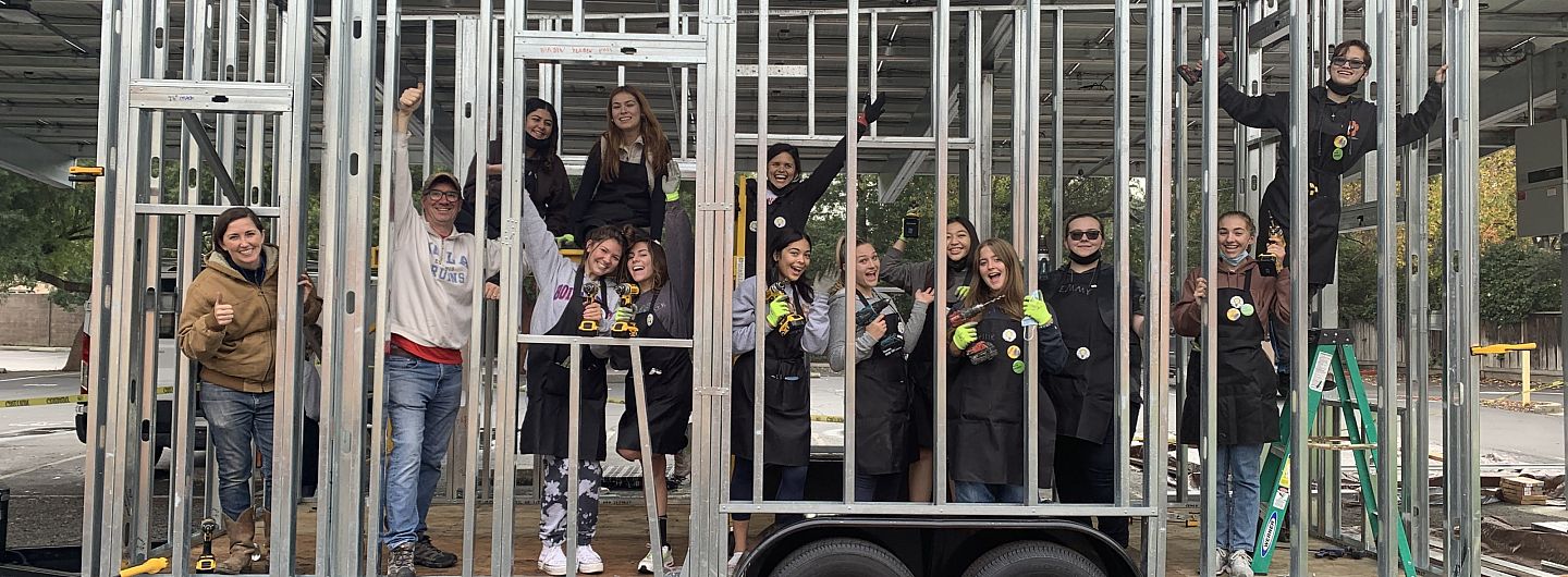 Twelve students and three adults smile widely inside the steel framing of the tiny house they're constructing.