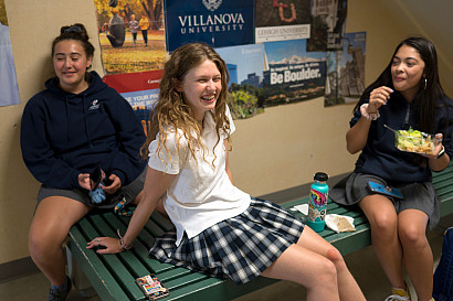Three students sit on a bench under the staircase